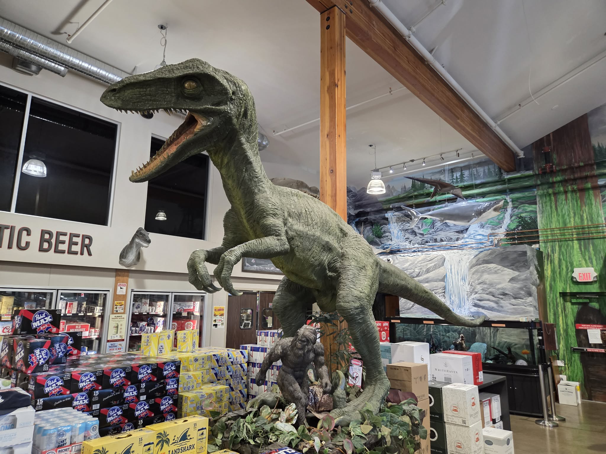 Velociraptor towering over a Bigfoot statue near the craft beer aisle, with the aquarium mural behind