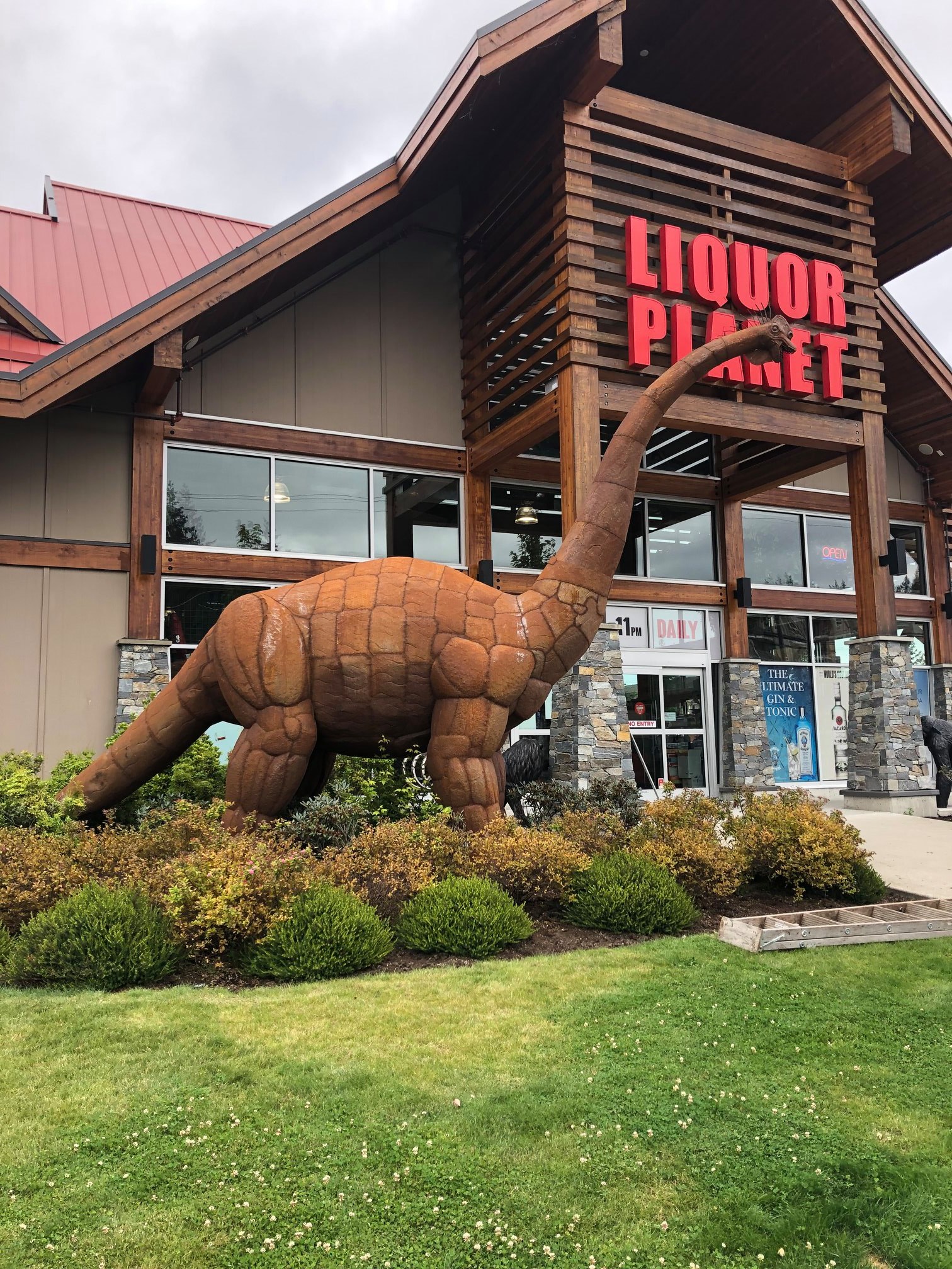 A rust-colored Brontosaurus statue out front of the Liquor Planet storefront, neck stretching up toward the Liquor Planet sign